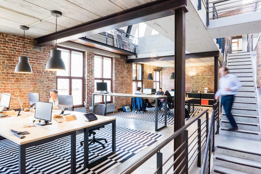 Office with an open floor plan, brick walls, and many computer screens and touch screens on desks.